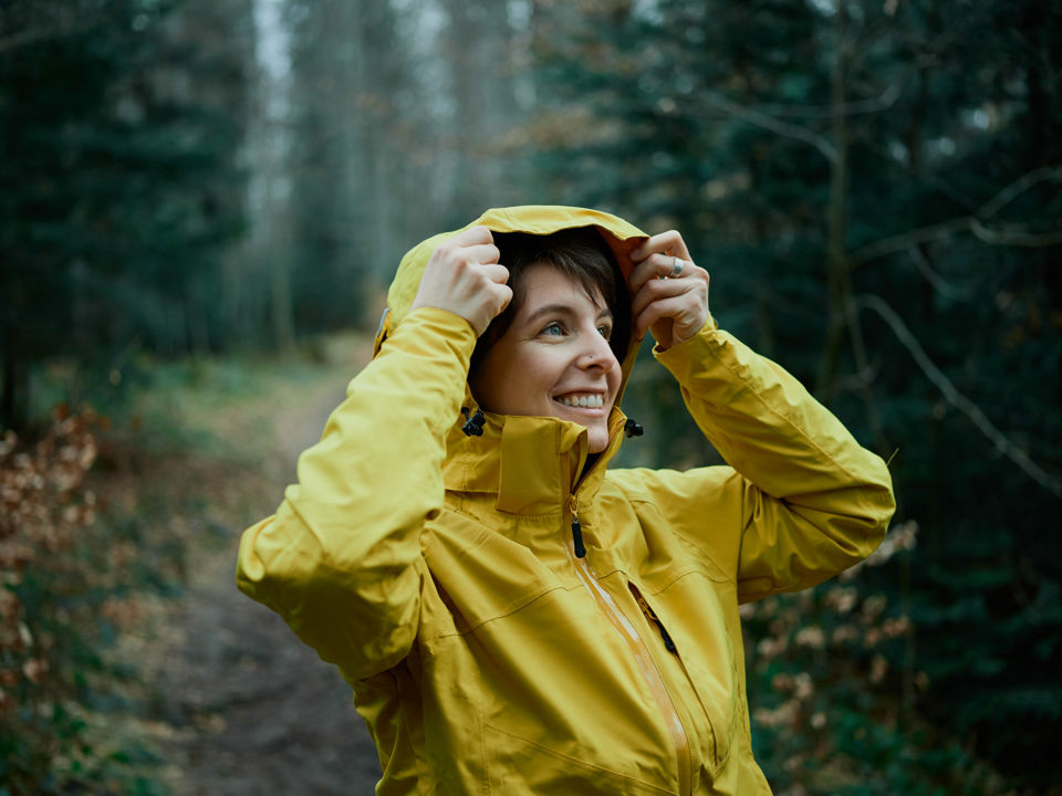 Eine glückliche Frau in gelber Regenjacke schaut sich beim Waldspaziergang etwas an
