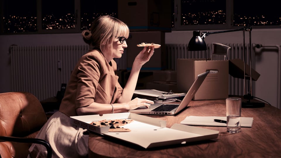 Eine Unternehmerin arbeitet spät am Abend im Arbeitszimmer und isst ein Stück Pizza vom Lieferdienst