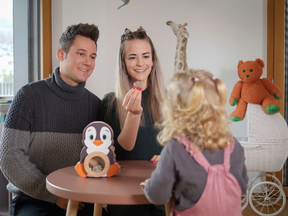 Junge Eltern geben ihrer kleinen Tochter im Kinderzimmer eine Münze für das Pinguinkässeli