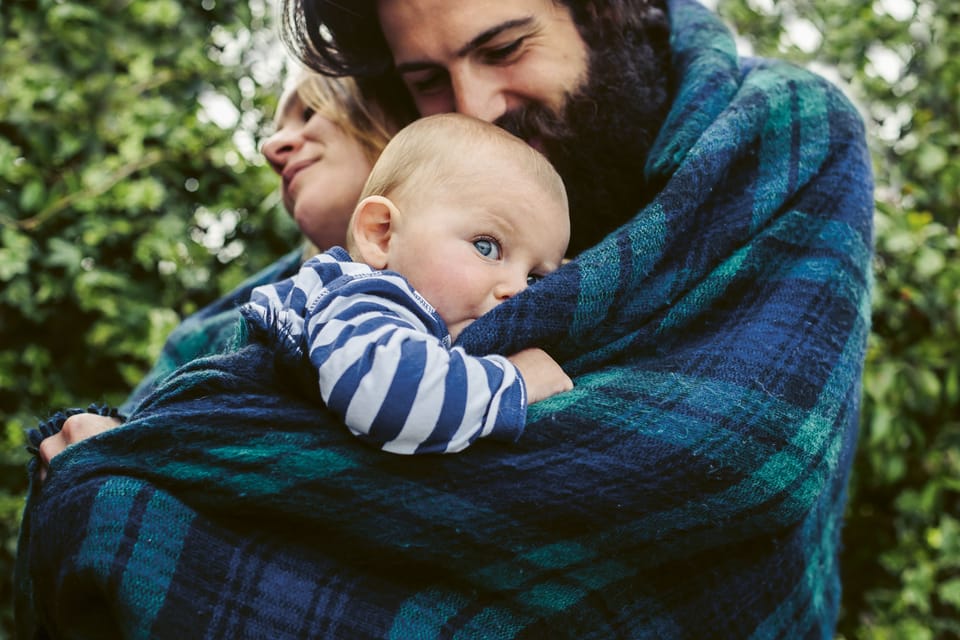 Frischgebackene Eltern kuscheln sich mit ihrem Baby im Grünen in eine warme Decke