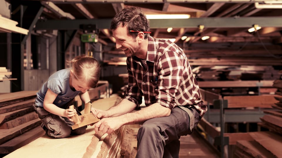 Ein Schreiner zeigt seiner kleinen Tochter in der Werkstatt  fröhlich den Umgang mit einem Hobel