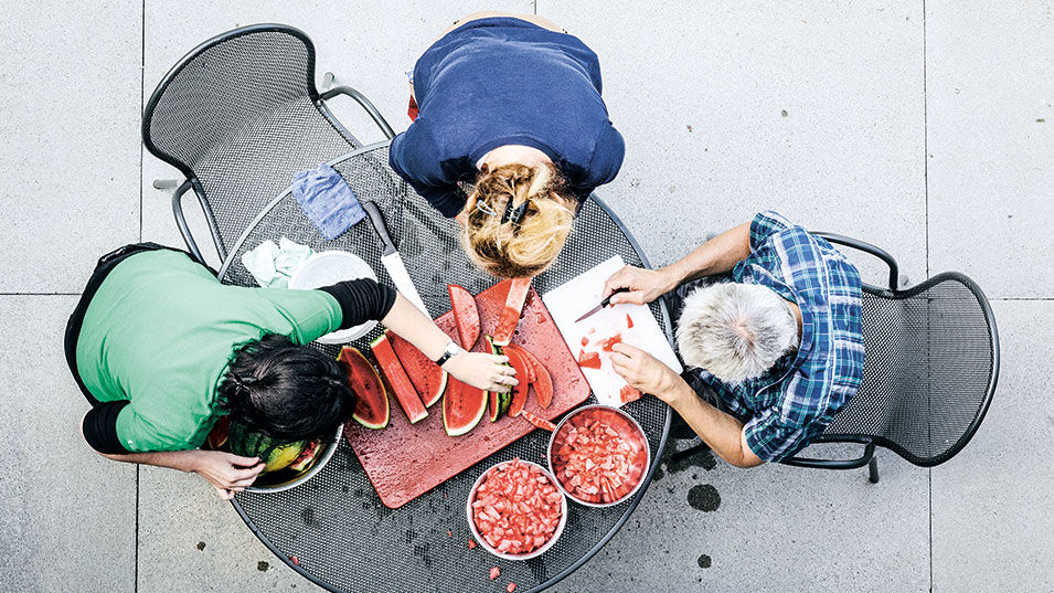 Blick auf drei Personen, die an einem Tisch Wassermelonen in Stücke schneiden